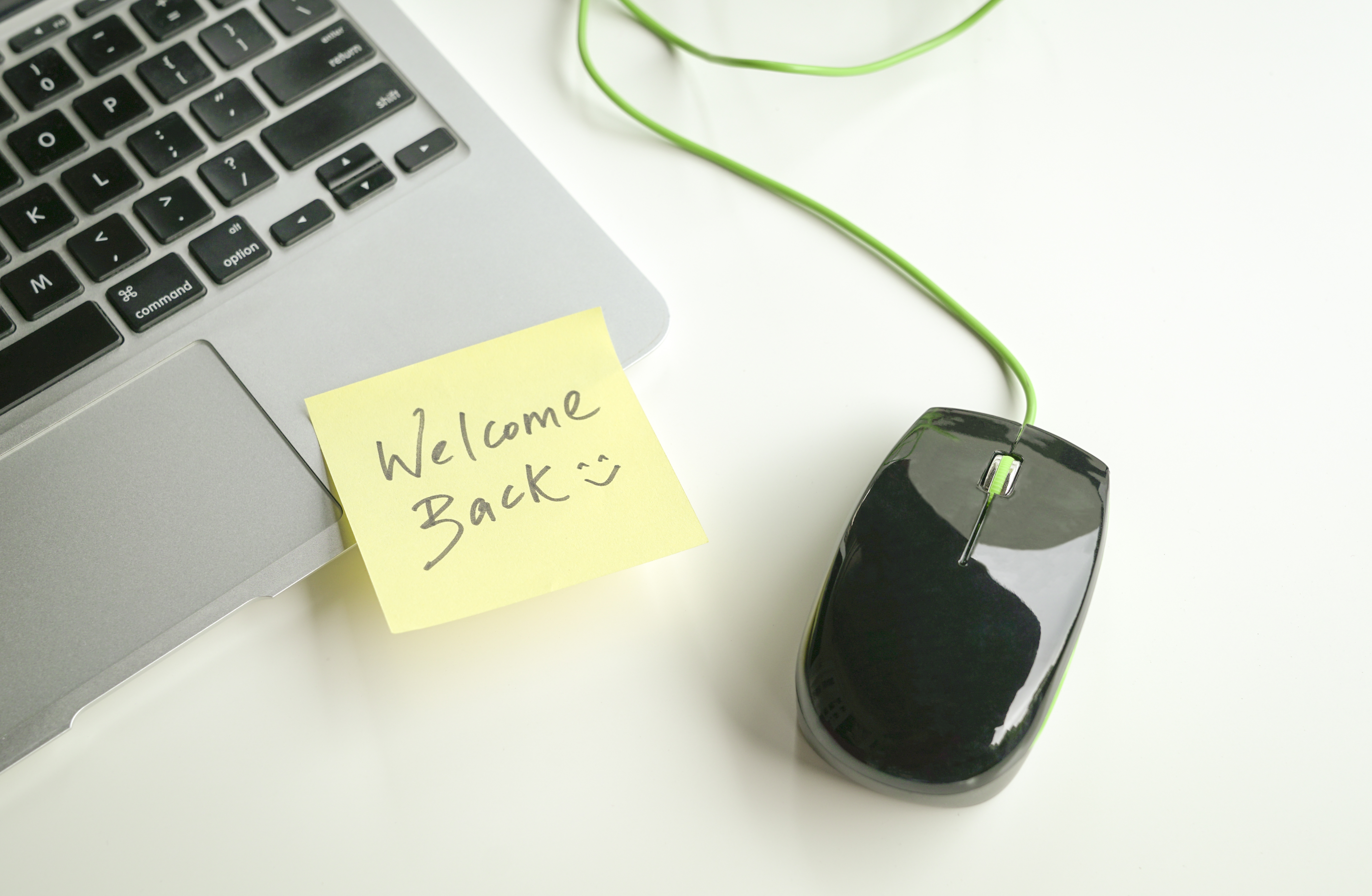 Laptop computer and wired mouse with welcome back and smiley face on a yellow note stuck to the keyboard