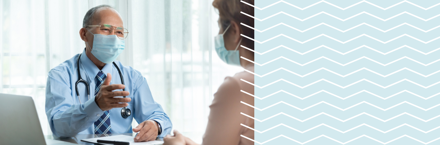 masked male physician sitting across a desk from a female patient wearing a mask