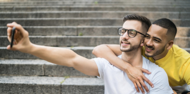 Two men taking a selfie on concrete steps. The man behind is wearing a yellow shirt with his arm around a man in a white shirt.