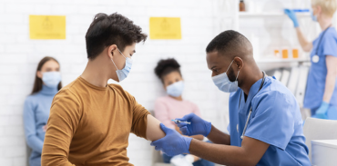 A young adult male receiving a vaccination from a man in blue scrubs and a mask