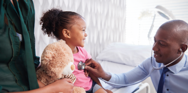 male physician on his knees while using a stethoscope on a young female patient sitting on a hospital bed with her teddy bear