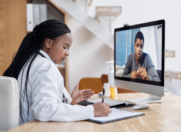 a black female physician sits infront of a computer providing virtal care to a paient on the computer monitor